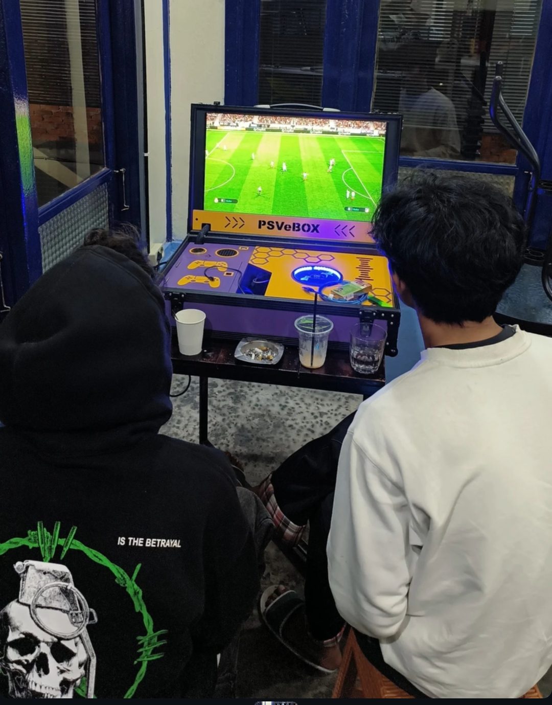 Unit PSVeBOX terbuka di meja kafe, dua orang bermain sepak bola PS4